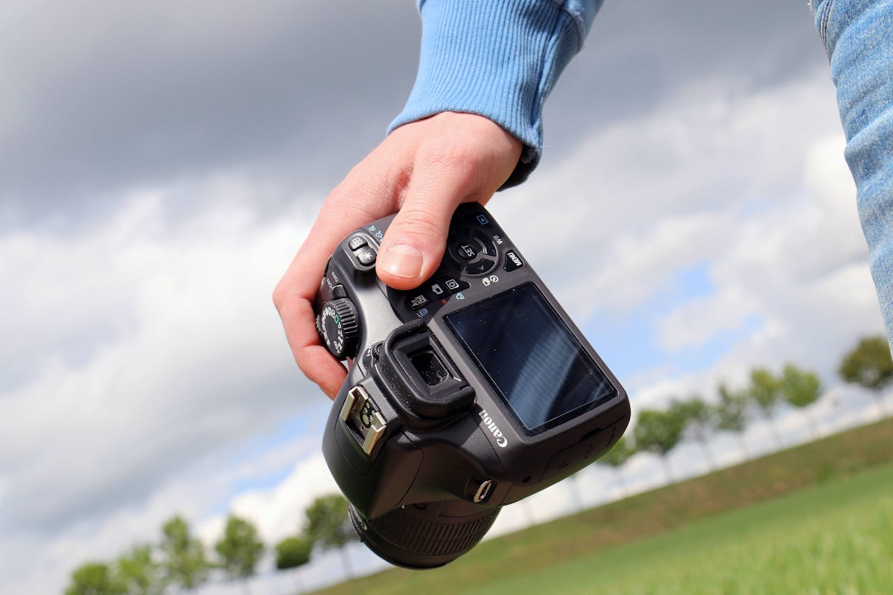 Person holding DSLR camera in hand, ready for outdoor photography against a cloudy sky.